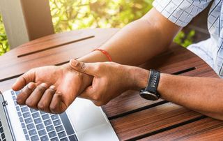 Person at a laptop gripping a painful wrist, a common sign of carpal tunnel syndrome treated with in-office WALANT release at Tyrance Orthopedics in Delray Beach.