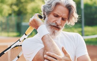 Older tennis player on a clay court holding his racket and gripping his painful elbow, a sign of tennis elbow treated with PRP at Tyrance Orthopedics in Delray Beach.