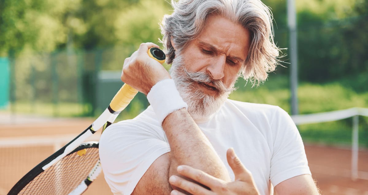 Older tennis player on a clay court holding his racket and gripping his painful elbow, a sign of tennis elbow treated with PRP at Tyrance Orthopedics in Delray Beach.