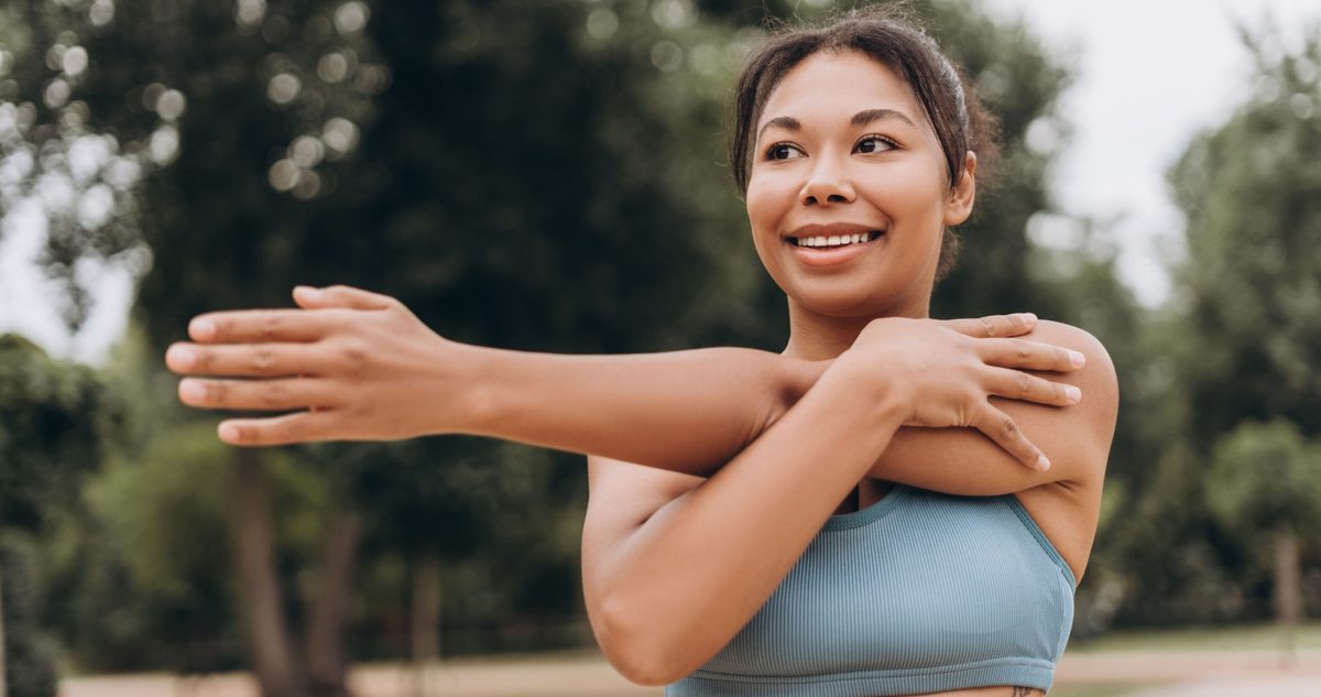 Woman easing into a cross-body shoulder stretch outdoors, the warm-up that keeps an irritated bursa from flaring.